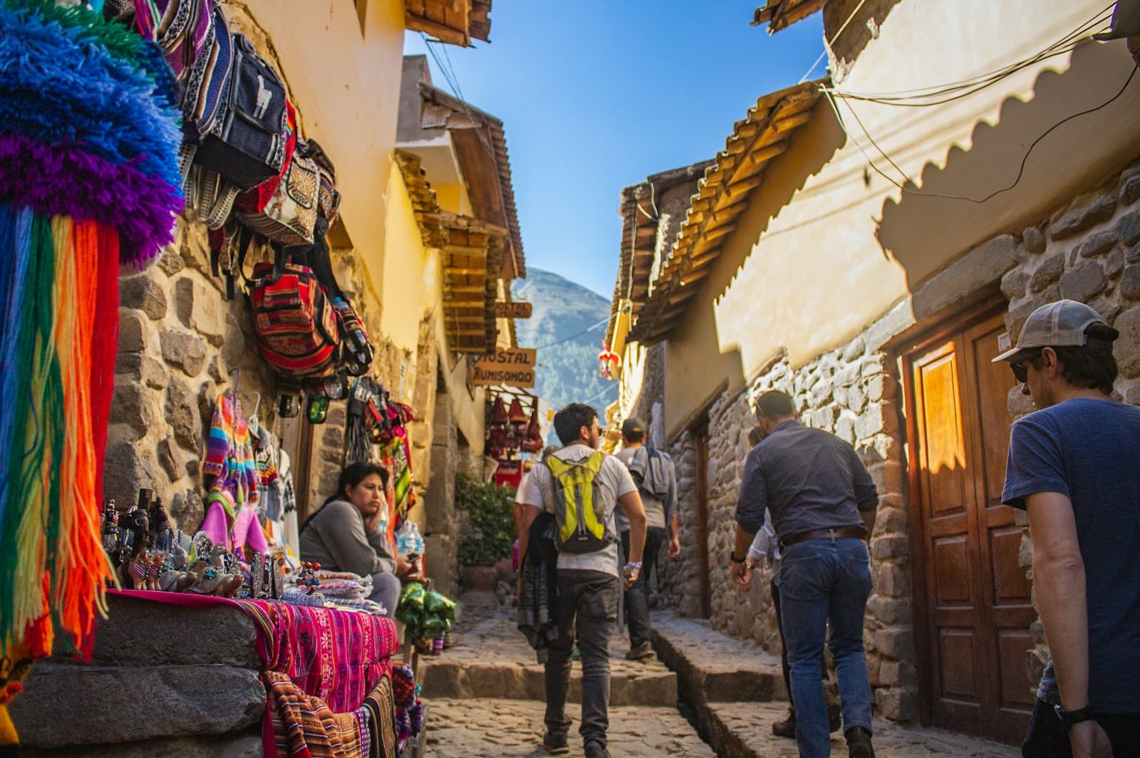 Viajeros con mochila caminando por una calle de piedra en Ollantaytambo, junto a puestos de tejidos andinos de colores.